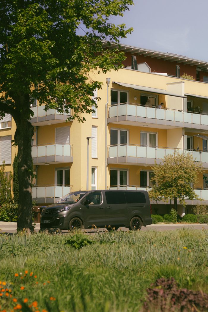 A serene urban scene featuring a beige building and a black van in a lush green park on a sunny day.